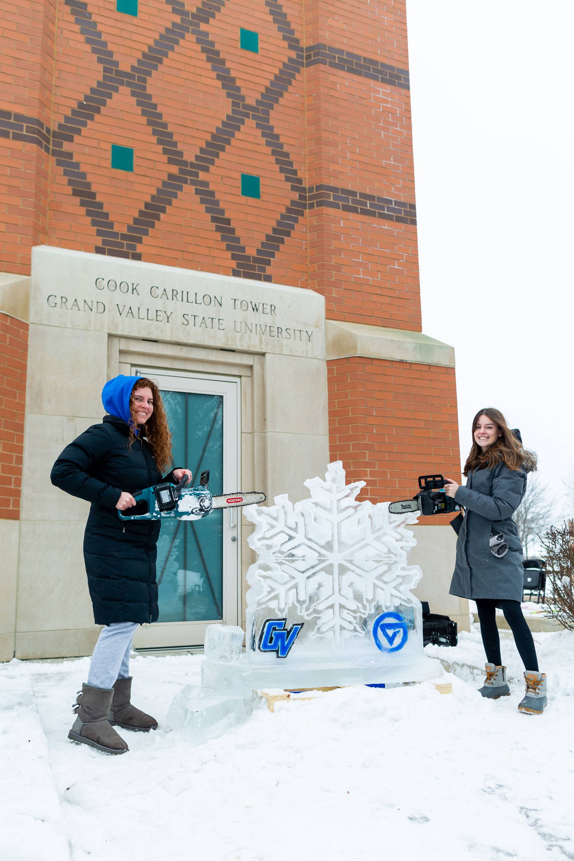 Two women posing with chainsaws by snowflake ice sculture